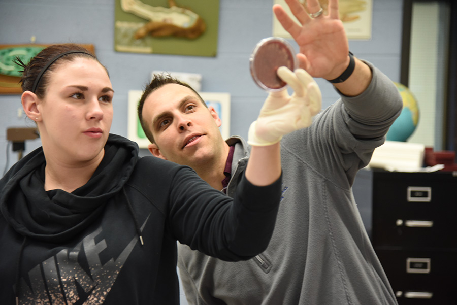Student wearing latex gloves examines petri dish at arm's length, with guidance from instructor.