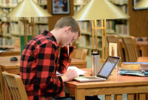 Photo of male student studying with laptop and books at one of our library desks with lamps