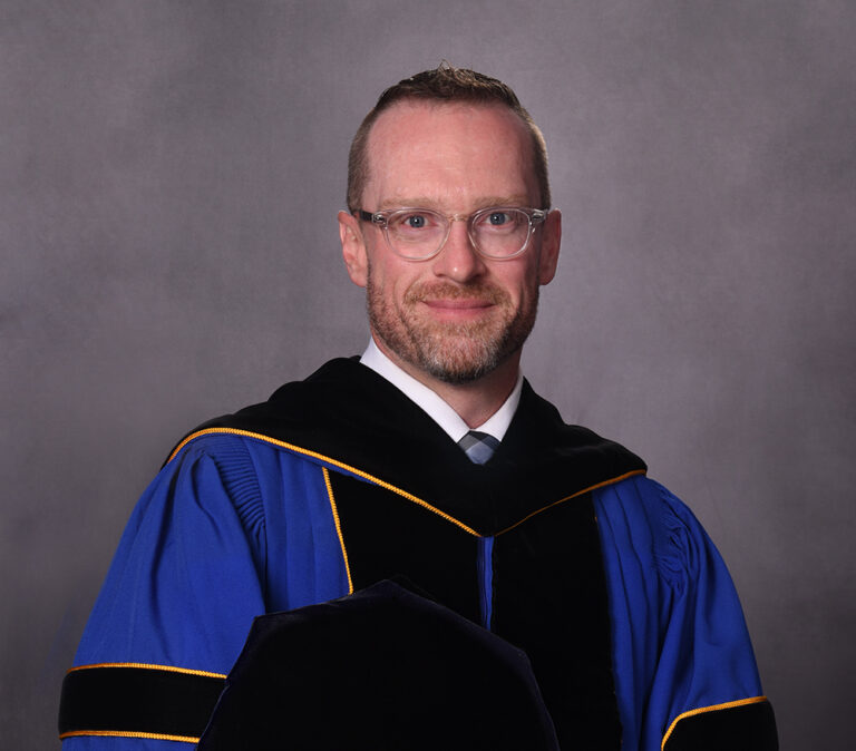 GCC President Doctor Craig Lamb, posing in academic regalia, with his cap held to his chest. His regalia are blue and black, with yellow piping.
