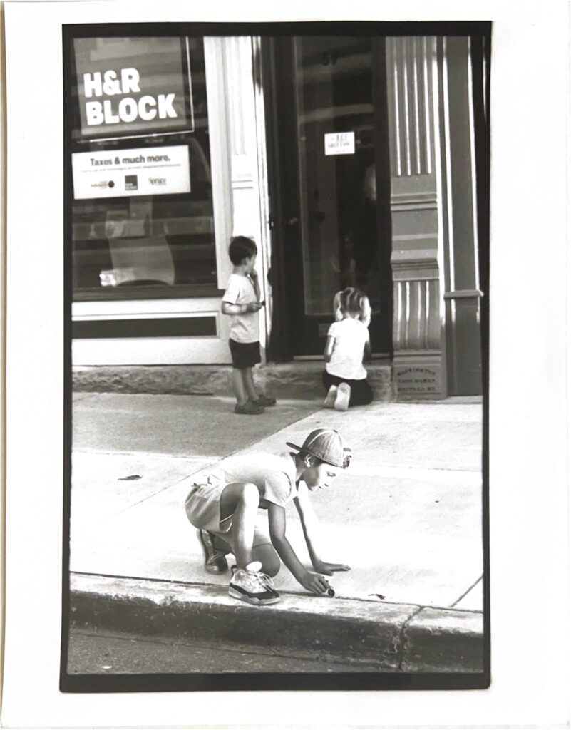Black and white photo of children playing on a village sidewalk in front of an H&R Block