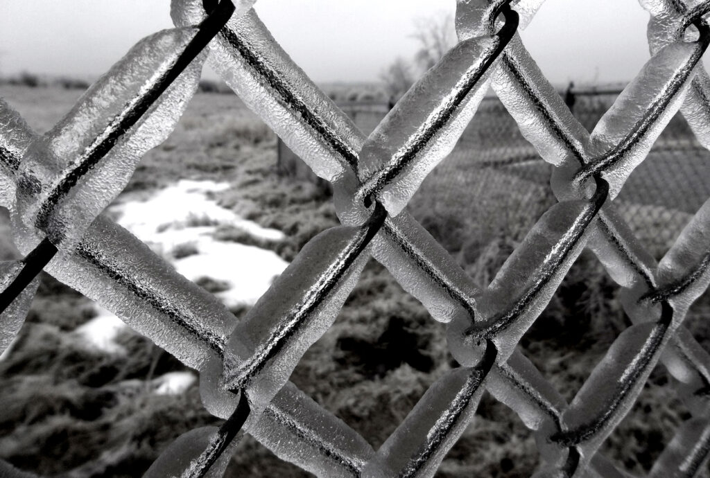 Black and white photo of ice covered fence with patchy snow in background