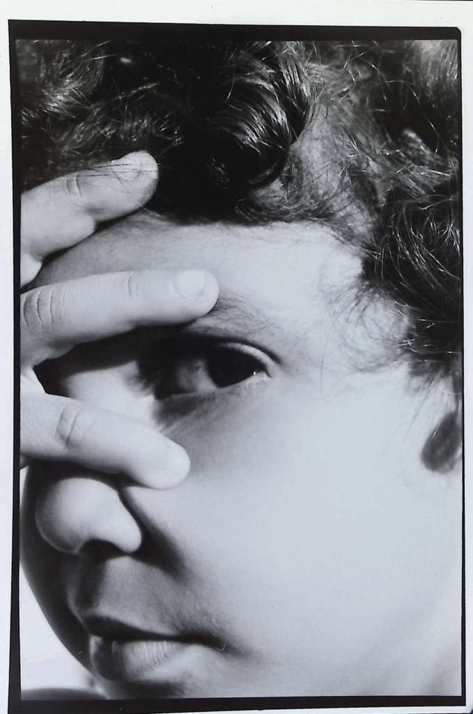 Black and white picture of child with hand spread over face, one eye looking at camera